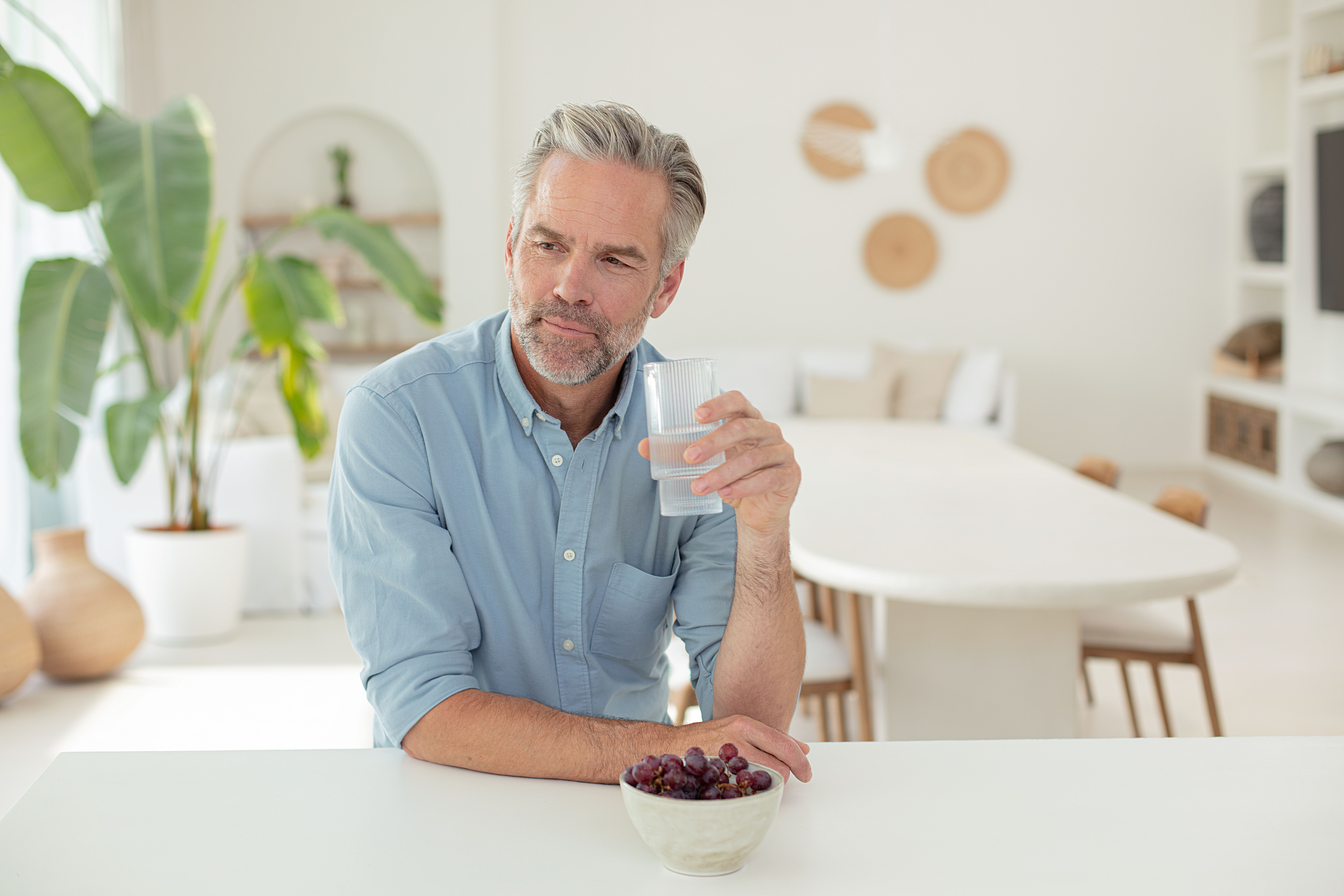 A mature man sits at a clean white counter, holding a glass of water and looking thoughtfully to the side, with a bowl of grapes nearby.